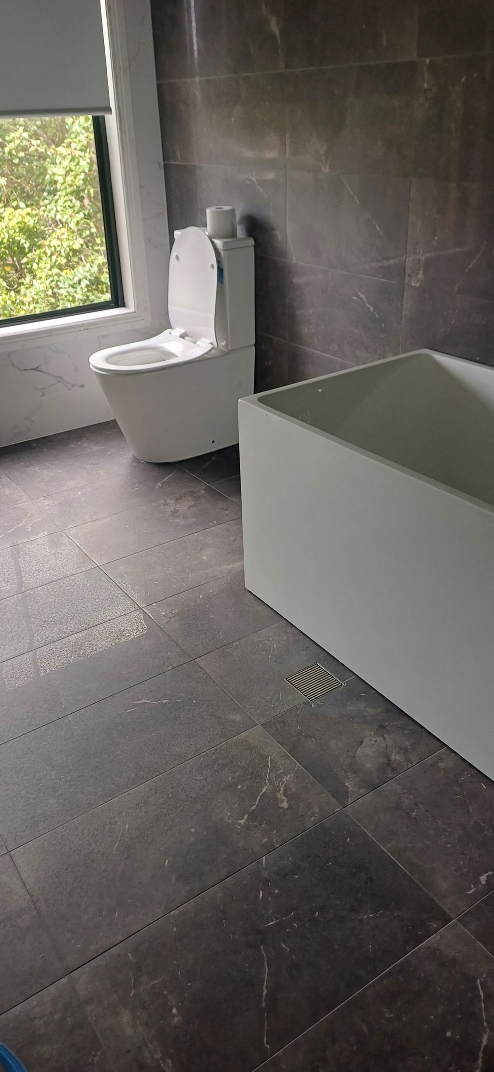 Bathroom with a dark stone-look tiled floor and walls, a freestanding bath and a square floor waste